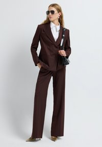 Mujer vestida con un traje de rayas marrones, camisa blanca, tacones dorados, bolso negro de hombro y gafas de sol tipo aviador, de pie contra un fondo liso.