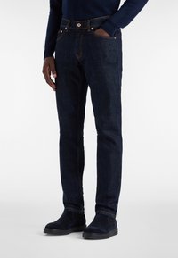 Moški v temno modrih slim-fit kavbojkah z oranžnim šitjem, mornarsko modrem puloverju in temno zamšastih Chelsea škornjih, z eno roko v žepu.