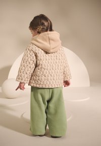 Veste matelassée beige à motif floral avec capuche beige, associée à un pantalon vert ample, enfant pointant du doigt, fond doux avec des formes géométriques.