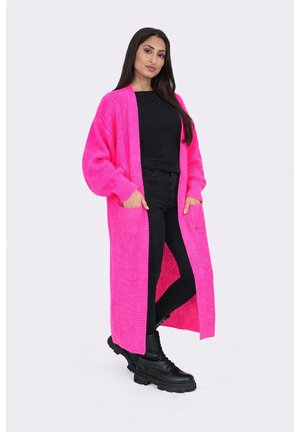 Kvinde står med hænderne i lommerne iført lyserød lang strikket cardigan, sort top, sorte bukser og sorte militærstøvler på hvid baggrund.