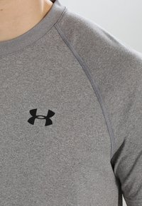 Detailní záběr osoby v šedém sportovním tričku s černým logem Under Armour na hrudi.