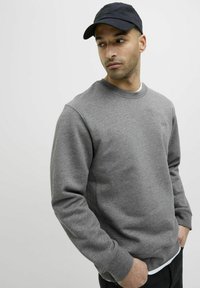 Felpa grigia con collo rotondo e polsini a coste; presenta un discreto dettaglio del logo. Indossata con un cappellino nero e una maglietta bianca sottostante, creando un look a strati.