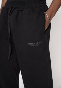 Pantaloni sportivi negri cu talie elastică și șnur. Au un logo discret în relief „MULTIPLY APPAREL” pe buzunarul din stânga.