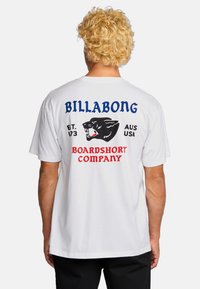 Persoon met krullend blond haar die een wit T-shirt draagt met een zwarte panter en de tekst "Billabong Boardshort Company" op de achterkant.
