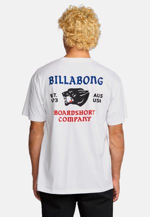 Person with curly blonde hair wearing a white T-shirt with a black panther and "Billabong Boardshort Company" text on the back.