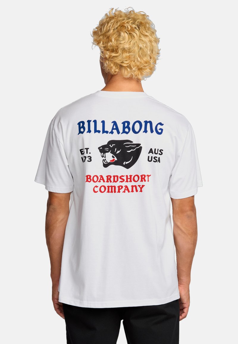 Persoon met krullend blond haar die een wit T-shirt draagt met een zwarte panter en de tekst "Billabong Boardshort Company" op de achterkant.