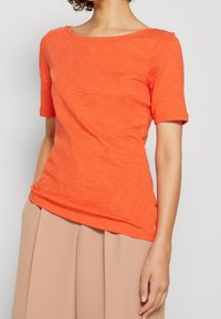 T-shirt arancione a maniche corte realizzato in tessuto texturizzato con scollatura ampia, abbinato a pantaloni beige a pieghe.