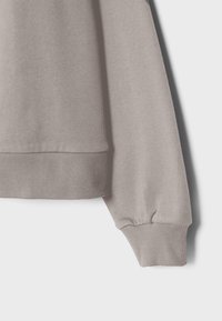 Lys beige strikket sweaterærme og nederste kant med ribbet manchet mod en hvid baggrund, der viser stoffets tekstur og syningsdetaljer.