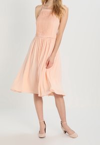 Robe halter de couleur pêche avec un bustier plissé, une jupe fluide et un tissu léger ; associée à des chaussures à talons hauts nude.