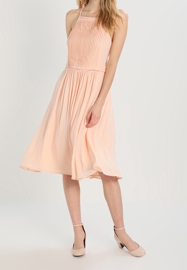 Robe halter de couleur pêche avec un bustier plissé, une jupe fluide et un tissu léger ; associée à des chaussures à talons hauts nude.