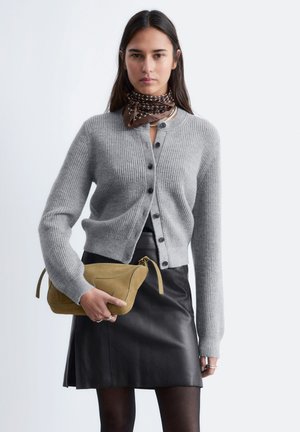 Woman wearing gray buttoned cardigan, black leather skirt, brown patterned scarf, holding a tan suede clutch bag.
