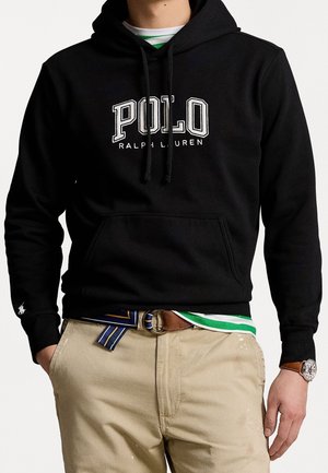 Sort hættetrøje med teksten "POLO Ralph Lauren", khakibukser med mønstret bælte, grøn- og hvidstribet skjorte under, armbåndsur på højre håndled.