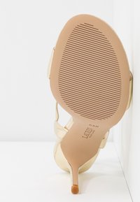 Sandale haute en or avec un dessus métallique lisse, un talon aiguille élancé et une semelle en caoutchouc texturé avec des rainures horizontales.