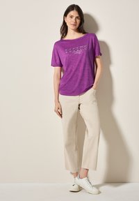 T-shirt violet à manches courtes avec un design imprimé ; associé à un pantalon beige clair ample et des baskets blanches. Texture douce, coupe décontractée.