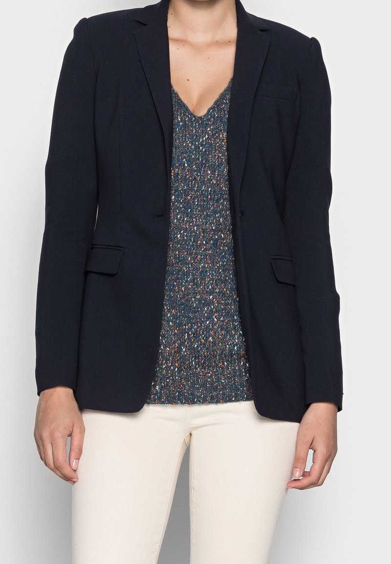 Femme portant un blazer noir sur un top en maille multicolore et un pantalon blanc, debout devant un fond uni clair.