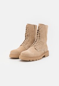 Un paio di stivali in suede beige con lacci, cerniere laterali e suole in gomma spessa, su sfondo bianco.