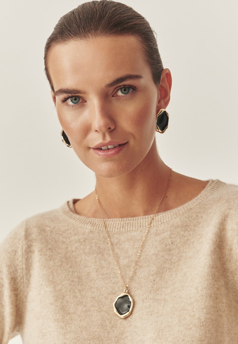 Černé šperky se šperkovým kamenem, které obsahují geometrický přívěsek na zlatém řetízku a odpovídající náušnice, v kombinaci se světle béžovým svetr.