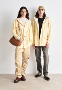 Jachete galbene de ploaie cu închidere prin buton, pantaloni cargo bej, o geantă de umăr maro și încălțăminte casual. Textură moale, croi lejer.