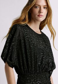 Robe noire à coupe ample et drapée, tissu texturé avec un motif de sequins, et fronces élastiques à la taille.