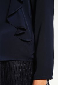 Blusa blu navy con un design con volant sulla parte frontale, maniche lunghe e un tessuto leggero e morbido. Abbinata a una gonna plissettata testurizzata.