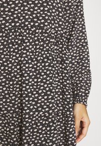 Robe noire avec motif de points blancs irréguliers, manches longues avec poignets froncés et une main reposant près de la couture latérale.