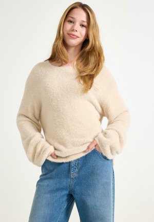 Beige flauschiger Pullover mit Rundhalsausschnitt und übergroßen Ärmeln, kombiniert mit blauen Jeans. Die weiche Textur und der lockere Schnitt sind bemerkenswerte Details.