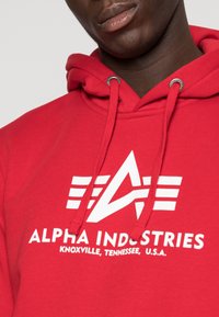 Punainen huppari, jossa on nauha ja hopeiset rei'itykset. Siinä on valkoinen logo, jossa on kolme vaaka-viivoitinta ja "ALPHA INDUSTRIES" -teksti.