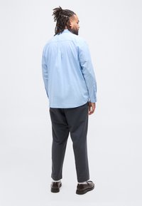 Άντρας με κοτσίδες, ντυμένος με ανοιχτό γαλάζιο πουκάμισο με μακριά μανίκια, σκούρο γκρι παντελόνι, άσπρες κάλτσες και καφέ μοκασίνια, στέκεται σε λευκό φόντο.