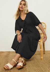 Vestido negro hasta la rodilla con una tela texturizada, mangas cortas abullonadas, escote en V y un detalle de lazo en el frente; combinado con sandalias beige.