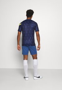 Maglia da calcio blu navy con motivo a pois, accentuata da spalline gialle, abbinata a pantaloni shorts blu, calze alte bianche e scarpe sportive nere.