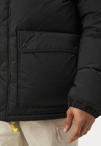 Black puffer jacket with a quilted texture, featuring a front pocket with snap closure and a drawstring hem for adjustable fit.