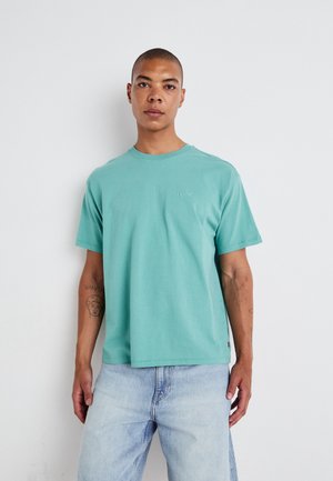 T-shirt turquoise à manches courtes avec un col rond, arborant un petit logo sur la poitrine gauche et une coupe décontractée. Associé à un jean bleu clair.