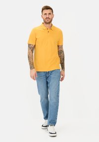 Geel poloshirt met een kraag, korte mouwen en een drieknopenrij. Draag het met lichtblauwe relaxed-fit jeans en witte sneakers.