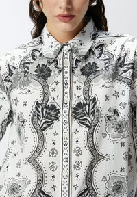 Camicia bianca con bottoni, caratterizzata da un motivo floreale e geometrico in nero e grigio, con colletto alla cinese e dettagli in pizzo lungo la parte anteriore.