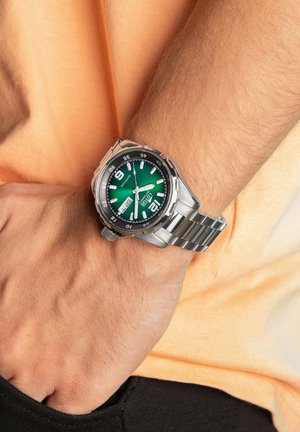 Silver metal watch with a green dial, marked with black numbers and a date function, worn on a wrist against a light orange shirt.