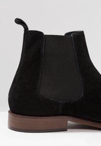 Bottine Chelsea en daim noir avec panneaux latéraux élastiques, bout arrondi et talon en bois. Le design présente une texture lisse et des coutures minimales.