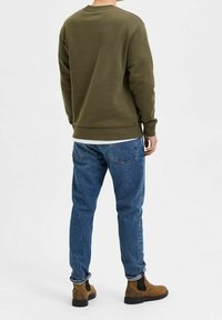 Olivově zelená mikina s dlouhými rukávy, pohled zezadu; v kombinaci s modrými džínami a hnědými kotníkovými botami s černými elastickými bočními panely.