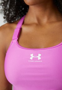 Rosa Under Armour Sport-BH mit verstellbaren Trägern, Rundhalsausschnitt, glatter Textur und einem weißen Logo auf der Vorderseite.