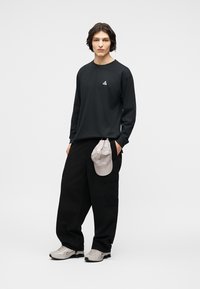 Νεαρός άνδρας που φοράει μακρυμάνικο μαύρο πουκάμισο, φαρδιά μαύρα παντελόνια με ένα ανοιχτό γκρι καπέλο να κρέμεται από την τσέπη, και ανοιχτό γκρι αθλητικά παπούτσια, όρθιος.