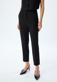 Zwarte op maat gemaakte broek met een hoge taille, plooien en taps toelopende benen, gemaakt van een gladde stof. Gepaard met glanzende zwarte hakken.
