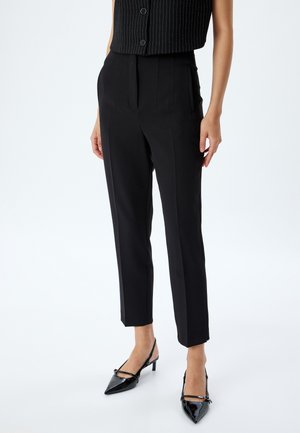Zwarte op maat gemaakte broek met een hoge taille, plooien en taps toelopende benen, gemaakt van een gladde stof. Gepaard met glanzende zwarte hakken.