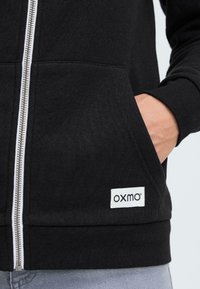 Černá mikina s kapucí na zip vyrobená z bavlněné směsi s bočními kapsami. Obsahuje stříbrný zip a bílou visačku se značkou "oxmo".