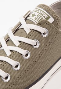 Baskets en toile vert olive avec lacets blancs, œillets en métal argenté et semelle en caoutchouc. Présente un logo Converse sur la languette.