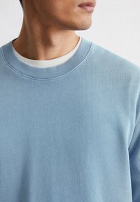Felpa azzurra chiara con colletto a girocollo a coste, caratterizzata da un tessuto texture e un collo interno bianco in contrasto. Dettaglio della parte superiore dell'indumento.