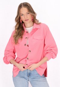 Camicia rosa a coste con maniche lunghe, chiusura con bottoni, taschino sul petto e polsini arrotolati abbinata a jeans di denim.