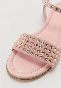 Pink sandal with flat sole, featuring a wide strap adorned with round, reflective studs. Smooth leather texture. Simple, open design.