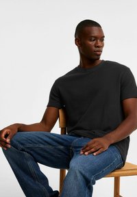 Sort tekstureret t-shirt med rund halsudskæring, korte ærmer og afslappet pasform, parret med blå denim jeans, mens der sidder på en træstol.