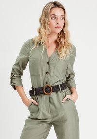 Olivgrüner Jumpsuit mit Knopfverschluss, zwei Brusttaschen und hochrollbaren Ärmel, kombiniert mit einem strukturierten schwarzen Gürtel mit einer Holzschnalle.
