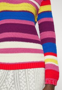 Randig tröja med flera färger som har zigzag-mönster i röd, blå, gul, rosa, lila och vit. Ribbstickade muddar och fåll.