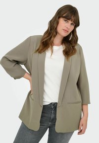 Blazer en verde oliva claro de tejido suave, con un diseño de frente abierto, mangas tres cuartos y dos bolsillos delanteros. Usado sobre una camisa blanca.